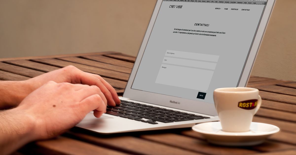 Mãos digitando em notebook exibindo um formulário de site, com xícara ao lado